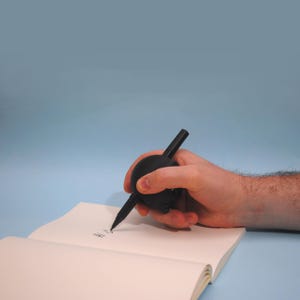 May include: A black pen with a round, black grip is being held by a hand and used to write on a blank notebook. The pen is designed to help with writing.