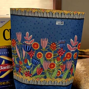 May include: A blue tote bag with a floral design featuring pink, red, and blue flowers. The bag has a black and white striped border and a yellow and black patterned top edge. Bagels and other food items are in the foreground.