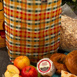 May include: A yellow, red, and green plaid reusable grocery bag with a small white label that says "Califia.com". The bag is filled with groceries, including apples, a round container of cheese labeled "Marin French Cheese Petite Truffle", and flatbreads.