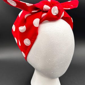 May include: A red headband with white polka dots, tied in a bow on top. The fabric is wrapped around a white foam head form, set against a black background. This accessory is a classic, retro style.