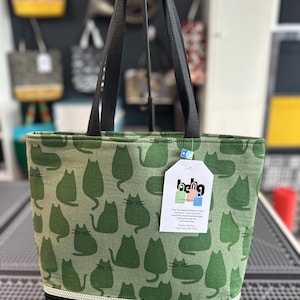 May include: Green tote bag with a repeating cat silhouette pattern. The bag has black handles and a black bottom with a white and gray patterned trim. A tag with the text "lache" is attached to the bag.