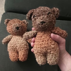 May include: Two handmade crochet teddy bears crafted from textured brown yarn. Each bear features small rounded ears, a dark stitched nose, and a simple embroidered mouth. The bears are similar in design but differ slightly in size and shade of brown.