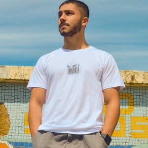 May include: A man wearing a white t-shirt with a black barcode graphic on the front. He is standing in front of a tiled wall and looking up.