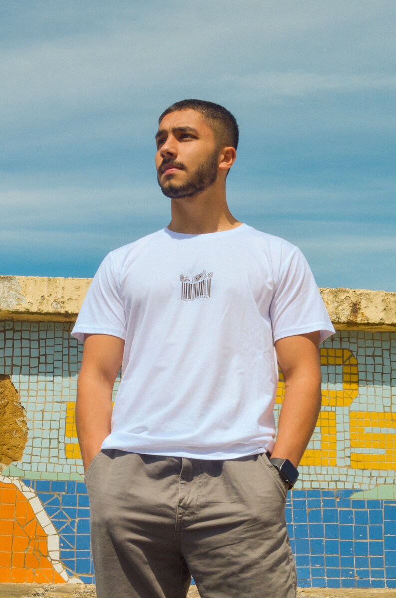 May include: A man wearing a white t-shirt with a black barcode graphic on the front. He is standing in front of a tiled wall and looking up.