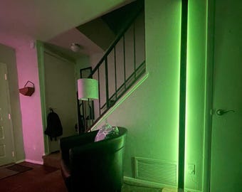 May include: A green LED floor lamp with a wooden base in a living room setting. The lamp is turned on and illuminating the room with a green glow.