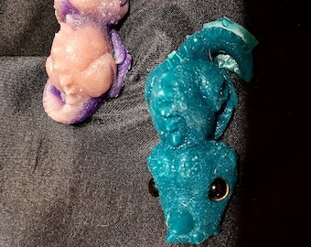 May include: Two small dragon figurines, one purple and one teal blue, with black eyes. The dragons are made of resin and have a textured surface.