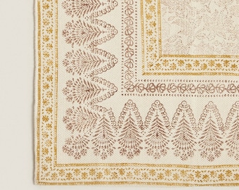 May include: A white cotton rug with a brown and gold floral pattern. The rug has a border with a repeating pattern of small flowers and leaves.