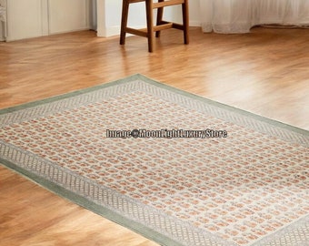 May include: A cream-colored rug with a green border and a repeating floral pattern. The rug is laid out on a wooden floor in a room with a wooden stool and a window with white curtains.