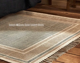 May include: A cream-colored woven rug with a geometric pattern in brown and black. The rug has a fringe border and is laid on a wooden floor.