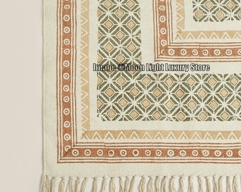 May include: A white cotton rug with a geometric pattern in shades of green, brown, and beige. The rug has a fringe border.