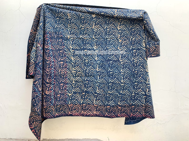 May include: A blue and white kantha quilt with a geometric pattern. The quilt is folded and hanging on a white wall.
