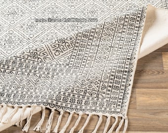 May include: A white and black patterned rug with fringe. The rug has a geometric design and is made of a soft, woven material.