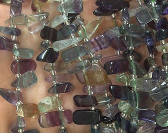Op de afbeelding: Strengen kleurrijke fluoriet edelsteen kralen. De kralen zijn een mix van paars, groen en helder, met onregelmatige vormen. De kralen zijn aan elkaar geregen, waardoor een ketting of armband ontstaat.