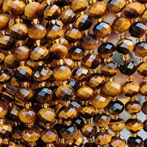 May include: Strands of faceted tiger eye gemstone beads in shades of brown and gold. The beads are strung together with gold-colored string, creating a shimmering effect. The beads are held in a hand.