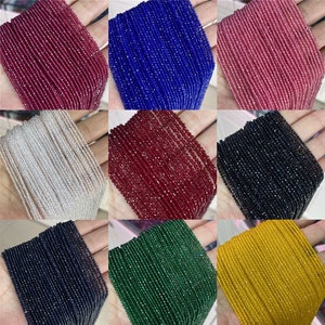 Może przedstawiać: Kolekcja sznurów fasetowanych koralików z kamieni szlachetnych w różnych kolorach, w tym czerwonym, niebieskim, białym, czarnym, zielonym i żółtym. Koraliki są małe i jednolite, tworząc gładki i spójny wygląd.