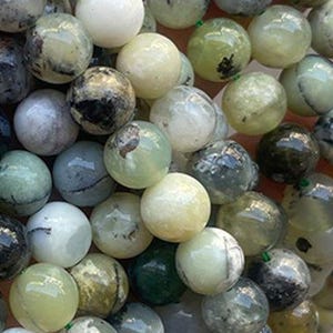 Puede incluir: Una colección de cuentas redondas de piedras preciosas en varios tonos de verde, blanco y gris. Las cuentas tienen una superficie pulida y lisa y están ensartadas, lo que sugiere que son para la fabricación de joyas. Las variaciones de color crean una estética natural y terrosa.