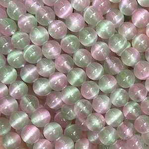 May include: Strands of round beads in shades of pink and green. The beads have a cat's eye effect, with a shimmering line running through each bead. The beads are strung together, creating a flowing, textured appearance. The beads are held in a hand.