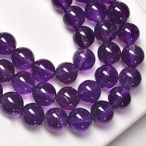 Puede incluir: Tres hilos de cuentas redondas de amatista pulidas en varios tonos de púrpura. Las cuentas están ensartadas, creando un collar o pulsera. Las cuentas se exhiben sobre una superficie blanca.