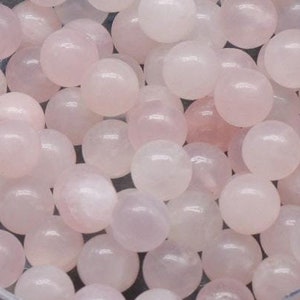 Può includere: Un contenitore di plastica trasparente pieno di numerose perle rotonde di quarzo rosa pallido. Le perle hanno un aspetto liscio e lucido e variano leggermente nella tonalità. Il contenitore è su una superficie grigio scuro.