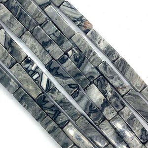 May include: Close-up of strands of rectangular beads. The beads are a mix of gray and white with a marbled pattern. The beads are strung together, creating a linear arrangement against a white background. Ideal for jewelry making.