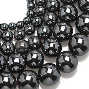 Puede incluir: Un conjunto de cuentas de hematites negras de varios tamaños, enhebradas en un hilo rojo. Las cuentas son redondas y lisas, con un alto pulido.