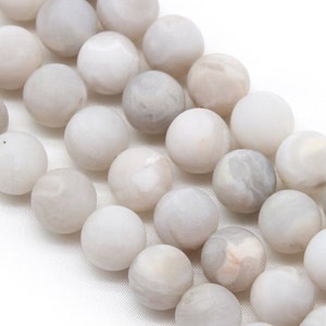 Peut inclure: Un brin de perles d'agate blanche avec des marbrures grises et brunes. Les perles sont rondes et lisses, et elles sont enfilées sur un fil transparent.