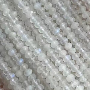 Puede incluir: Una hebra de cuentas de piedra lunar azul blanca. Las cuentas son redondas y tienen un acabado liso y pulido. Las cuentas están enhebradas en un hilo transparente. El texto "Blue Moonstone" es visible en la imagen.