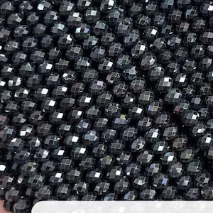 Puede incluir: Una hebra de cuentas de espinela negra, facetadas y redondas, con un acabado brillante y metálico. Las cuentas están enhebradas en un hilo transparente. El texto "Black Spinel" es visible en una etiqueta blanca.