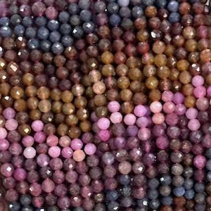 May include: Strands of faceted gemstone beads in shades of purple, pink, brown, and blue. The beads are small and strung together, creating a textured and colorful display. The gemstones are held in a hand.