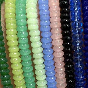 Peut inclure: Une collection de douze brins individuels de petites perles rondes et lisses dans une variété de couleurs, notamment blanc, vert foncé, vert lime, menthe, bleu clair, rose pâle, noir, bleu roi, transparent, crème et rouge foncé, disposés côte à côte.