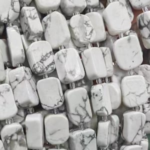 Könnte beinhalten: Eine Strähne weißer Howlith-Perlen mit quadratischer Form und grauer Maserung. Die Perlen sind auf einer durchsichtigen Gummikordel aufgefädelt.