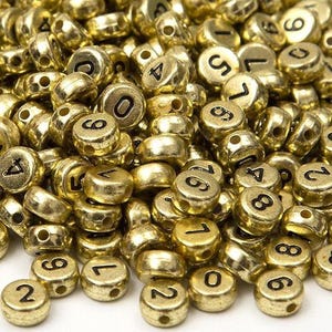 May include: A large pile of small, round, metallic gold-colored beads. Each bead features a single black numeral printed on its surface. The beads are scattered across a plain white surface, showing various numbers including 0 through 9.