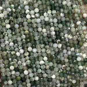 Op de afbeelding: Slierten kleine, gefacetteerde kralen in groen- en wittinten. De kralen worden in een hand gehouden, waardoor hun delicate formaat en kleurvariaties zichtbaar worden. De kralen zijn geregen, klaar voor het maken van sieraden.