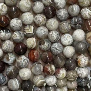 May include: Strands of round gemstone beads in shades of brown, gray, and white. The beads have a marbled appearance, with varying patterns and colors. The beads are held in a hand, showcasing their size and texture. The beads are approximately 8mm in diameter.