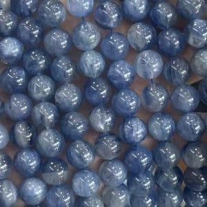 May include: Strands of round, blue gemstone beads. The beads have a marbled appearance with varying shades of blue and gray. The beads are held in a hand, showcasing their size and texture. These beads are suitable for jewelry making.