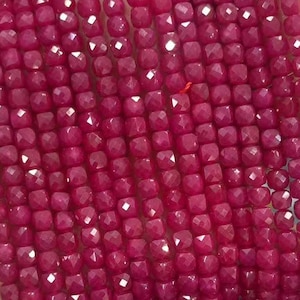 May include: A collection of deep magenta ruby beads, square-cut and faceted, held in a hand. The beads' polished surfaces reflect light, highlighting their consistent shape and size. These beads are suitable for crafting jewellery.