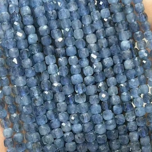 Puede incluir: Cadenas de gemas azules facetadas, cortadas en forma cuadrada. Las cuentas son de un color azul claro y parecen estar ensartadas. La imagen muestra las cuentas sostenidas en una mano, con un fondo borroso.