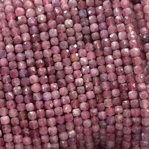 Op de afbeelding: Strengen gefacetteerde, vierkant geslepen, roze edelstenen. De kralen hebben een dieproze kleur en lijken aan elkaar geregen te zijn. De kralen worden in een hand gehouden, de achtergrond is wazig.