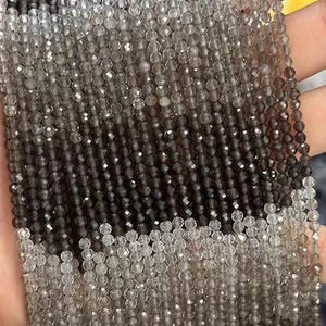 May include: Strands of faceted beads in a gradient of colors, from dark brown to clear. The beads are small and strung together, creating a textured surface. The beads are held in a hand, showcasing the color variations.