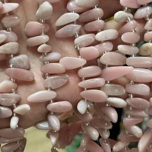 May include: Strands of pink opal gemstone beads. The beads are a mix of light and medium pink, with an irregular, oblong shape. The beads are strung together on a clear string, creating a flowing, organic look. The background is blurred, with hints of other colors.