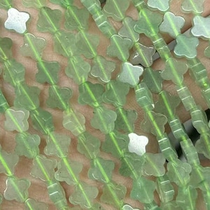Puede incluir: Primer plano de hebras de cuentas verde claro con forma de trébol y detalles blancos. Las cuentas están ensartadas, creando un patrón delicado. Las cuentas se sostienen en una mano, mostrando su color y diseño.