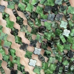 Puede incluir: Primer plano de hebras de cuentas verdes y negras en forma de trébol. Las cuentas están ensartadas, creando un patrón detallado. Las cuentas se sostienen en una mano, mostrando las variaciones de color y el diseño de las cuentas de piedras preciosas.