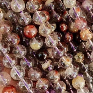 Puede incluir: Una hebra de cuentas redondas, translúcidas con inclusiones de morado, rojo y marrón. Las cuentas están enhebradas en un cordón elástico transparente.