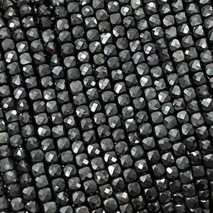 Op de afbeelding: Een streng zwarte gefacetteerde edelsteen kralen. De kralen zijn klein en vierkant en geregen op een doorzichtige draad. De kralen zijn gerangschikt in een nette, gelijkmatige rij.