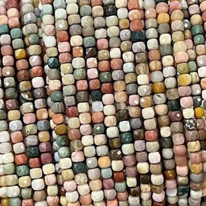 Puede incluir: Una hebra de cuentas de piedras preciosas multicolores, cada cuenta es un pequeño cubo facetado. Las cuentas son una mezcla de marrón, rosa, verde y blanco.