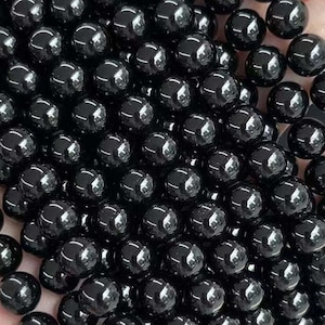 Può includere: Primo piano di fili di perline di ossidiana nera lucida e rotonda. Le perline sono di un nero intenso e lucido. Il testo in basso recita "A Grade Pure Black Obsidian" e caratteri cinesi.