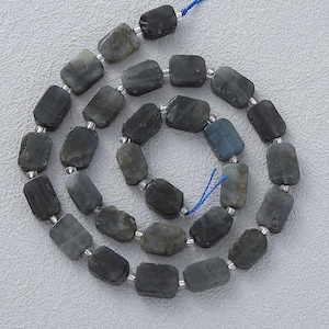 May include: A string of rectangular, dark grey beads with a subtle blue sheen. The beads are strung together with small silver-coloured spacers and a blue string, creating a necklace or bracelet.