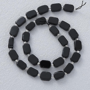 Può includere: Una collana di perline nere rettangolari. Ogni perla è liscia e lucida, infilata con un filo sottile e trasparente. Le perle sono uniformemente distanziate, creando un motivo coerente. Il design è semplice ed elegante, adatto alla creazione di gioielli.