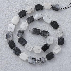 Puede incluir: Un collar de cuentas rectangulares en tonos negro, blanco y transparente. Las cuentas están ensartadas con un hilo negro, creando un patrón en espiral. Las cuentas tienen un aspecto moteado.