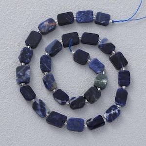 Puede incluir: Un collar de cuentas rectangulares de sodalita en tonos azules y blancos. Las cuentas están ensartadas en un cordón azul, con pequeños espaciadores plateados entre cada cuenta. Las cuentas miden aproximadamente 1,3 cm de largo.